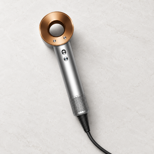 Dyson Supersonic HD-07 hair dryer in Nickel and Copper, staged on pale stone.